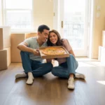 Couple eating pizza on floor of new apartment in El Mirage, AZ, surrounded by moving boxes and basic furniture.