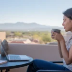 Woman drinking coffee on Tempe apartment balcony at sunrise, with laptop and plant on table