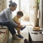 Parent helping child tie shoes in living room, budgeting app on table